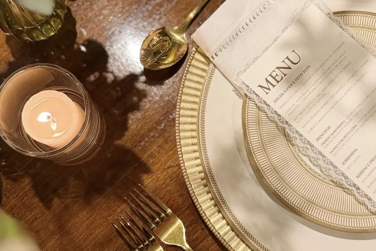 Detalhe de lugar posto sobre madeira escura: sousplat dourado trabalhado, prato branco com o menu impresso por cima, talheres dourados e copo de vela aceso ao lado