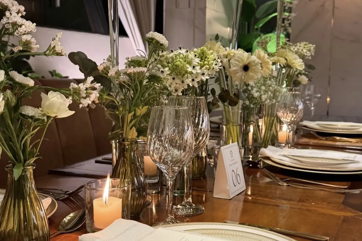Mesa longa posta à noite, com taças de cristal, talheres, vasinhos de flores brancas, velas acesas e o número da mesa em pé