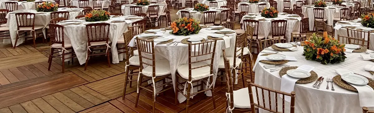Vista ampla de pavilhão montado para evento corporativo, com mesas redondas de toalha branca em série, cadeiras douradas e de madeira escura e arranjos de flores laranja ao centro de cada mesa, sobre piso de madeira