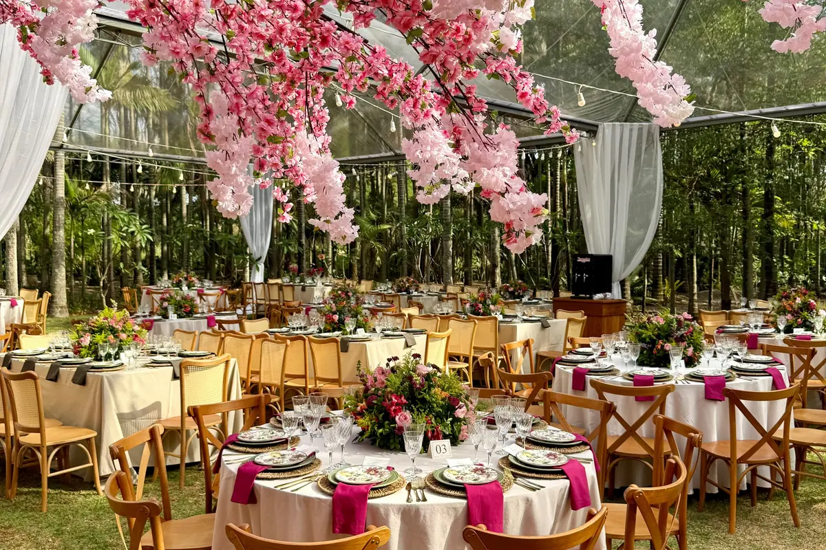 Festa montada no jardim, com mesas postas, cadeiras de madeira e arranjos de flores