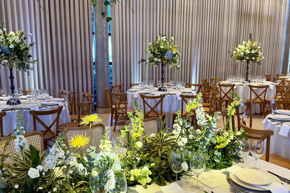 Mesa longa montada pela Totonho em salão, com caminho de flores brancas e amarelas no centro, e mesas redondas de toalha branca com cadeiras de madeira ao fundo