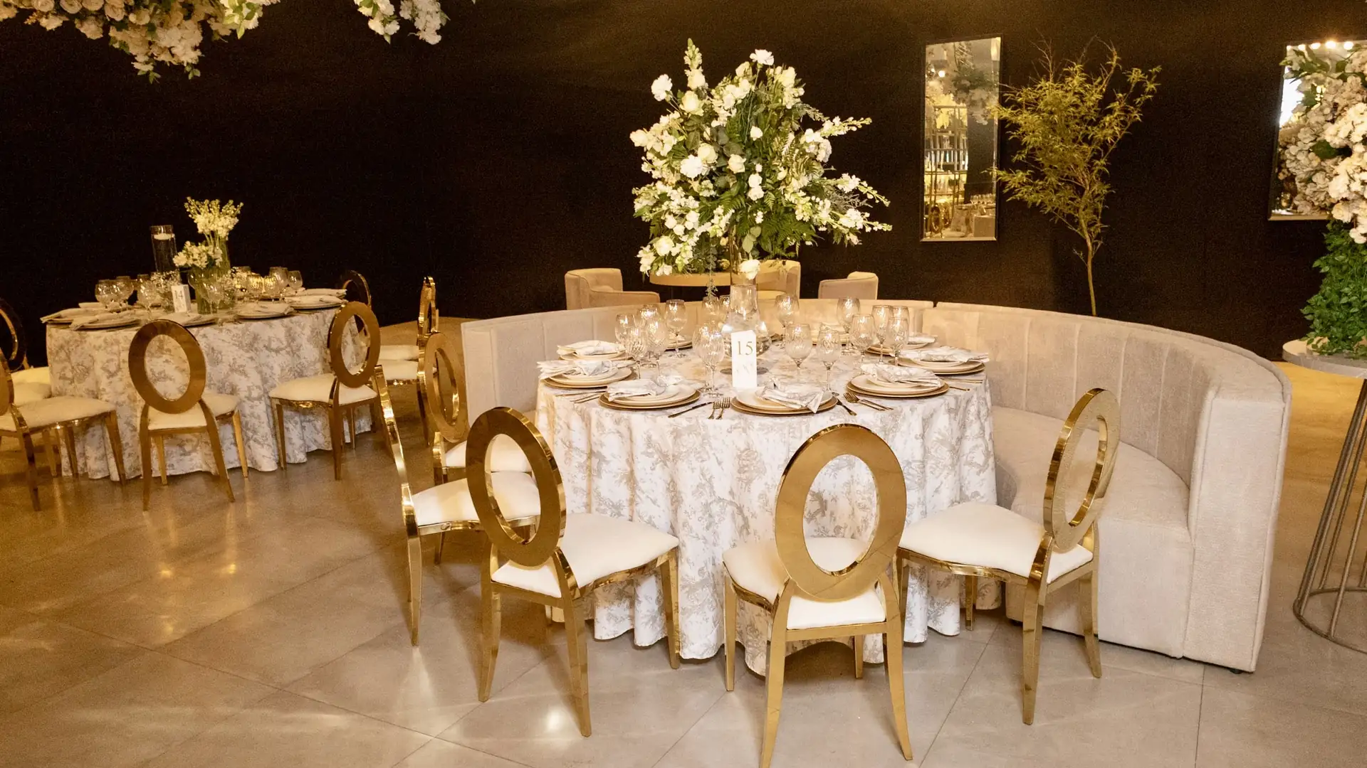 Salão montado à noite, com mesa redonda posta: toalha clara, sousplats dourados, taças de cristal, arranjo alto de flores brancas no centro e cadeiras douradas de encosto oval em volta