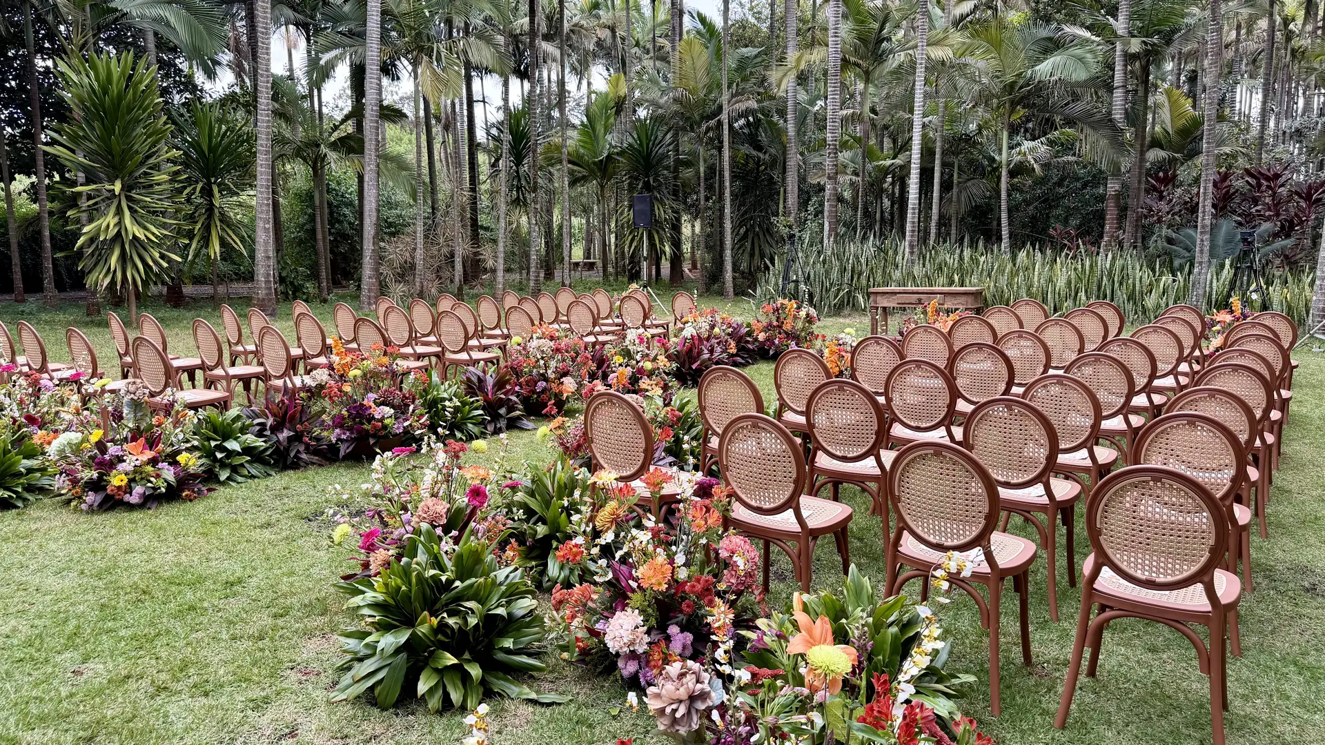 Cerimônia montada no gramado, com cadeiras de madeira de encosto oval em palhinha em duas alas e arranjos de flores tropicais coloridas no chão, entre palmeiras