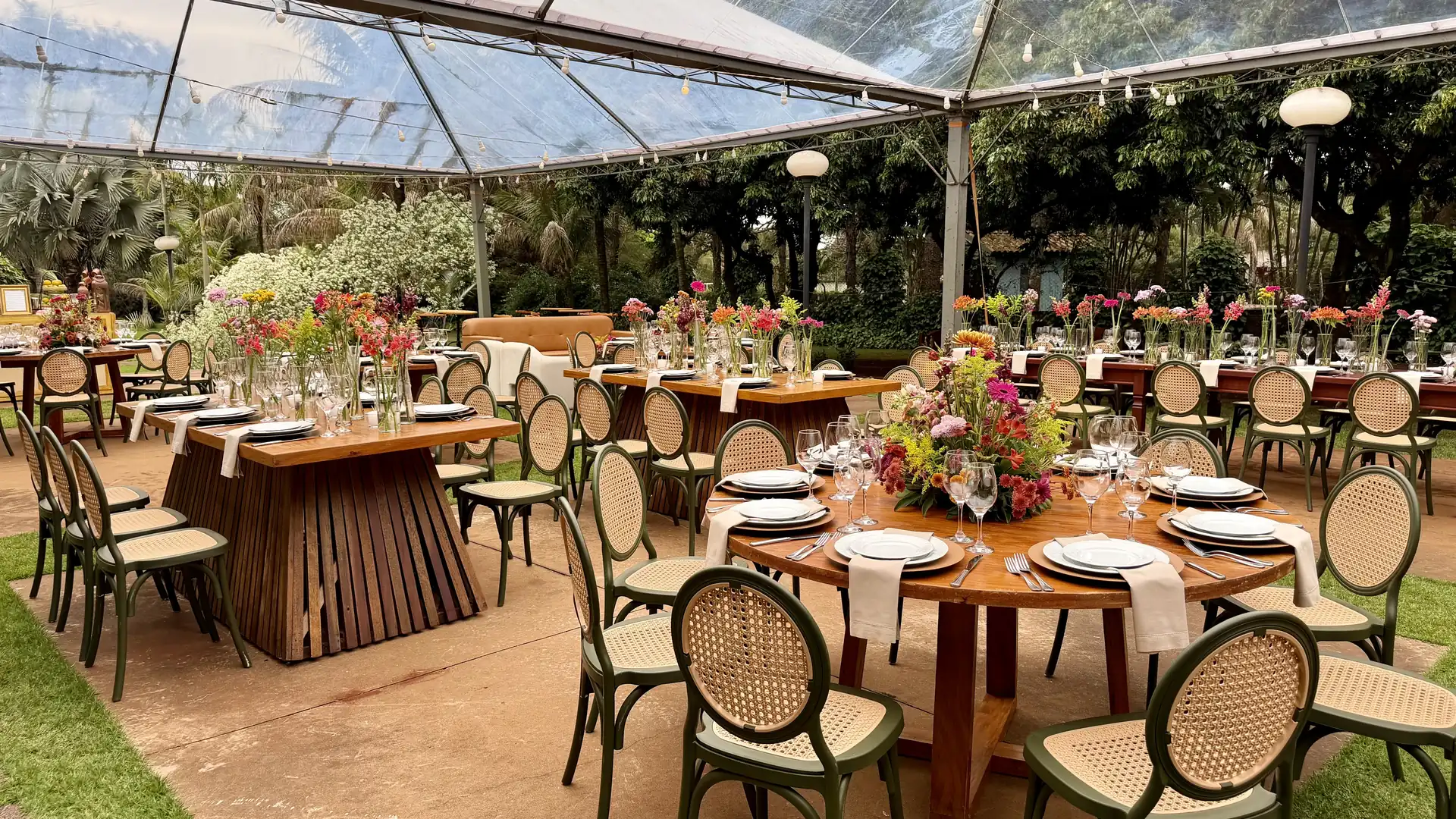 Recepção de dia sob tenda transparente, com mesas redondas e retangulares de madeira postas, cadeiras de encosto oval em palhinha e vasinhos de flores coloridas ao longo das mesas