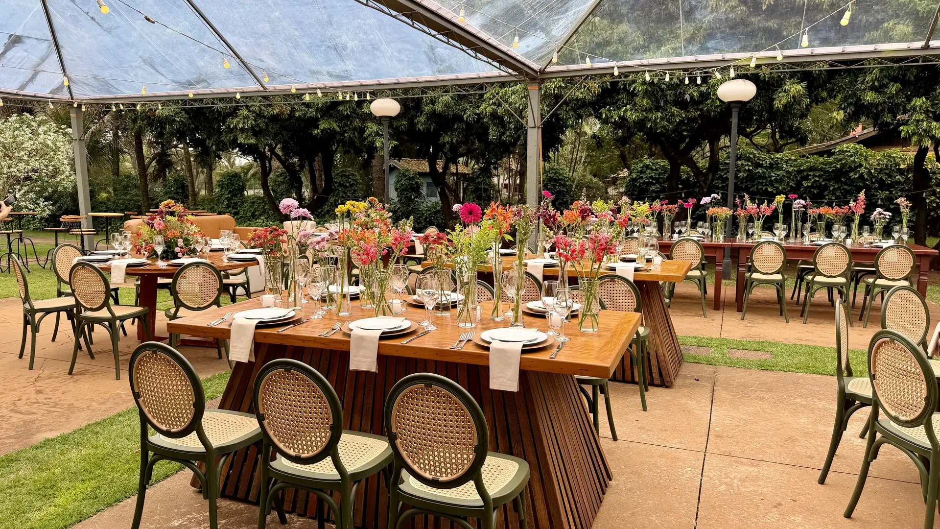 Mesas longas de madeira montadas sob tenda transparente no jardim, com fila de vasinhos de flores coloridas, guardanapos claros, taças e cadeiras de palhinha