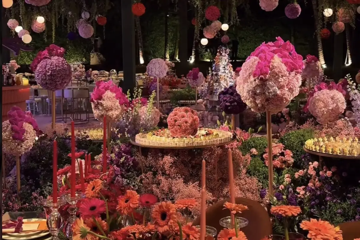 Mesa de doces montada com arranjos altos de flores em tons de rosa e laranja, boleiras e suportes redondos com doces, sob luminárias de papel penduradas