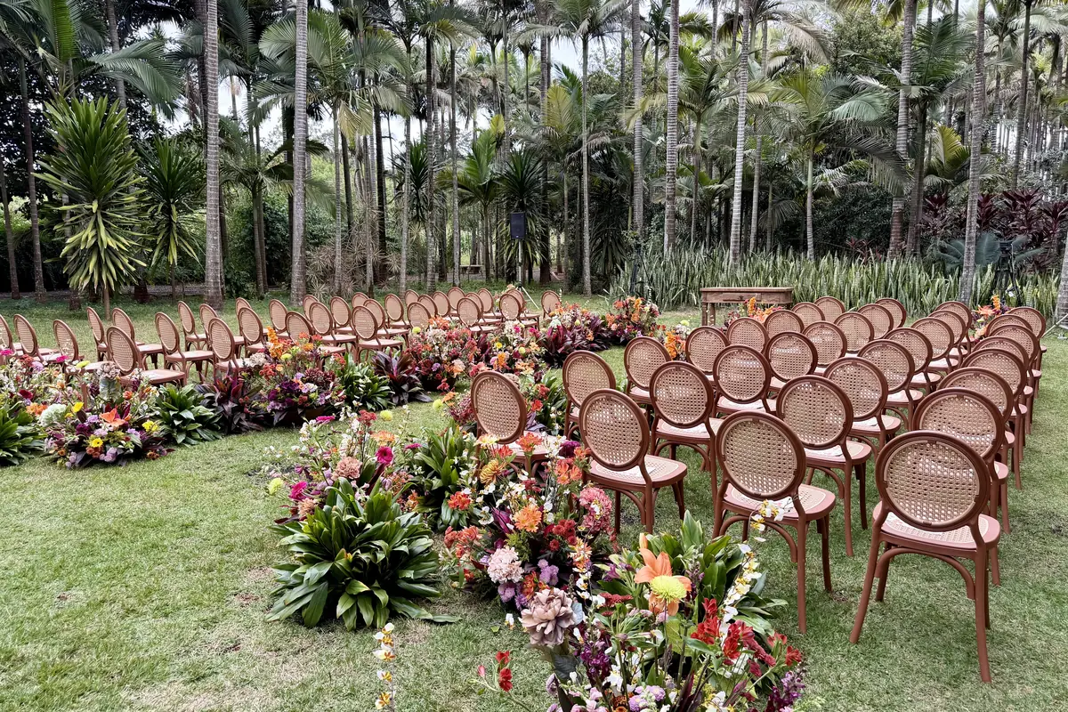 Cerimônia de casamento montada no gramado, com cadeiras de madeira de encosto oval em palhinha em duas alas e canteiros de flores tropicais coloridas ao longo do corredor, entre palmeiras