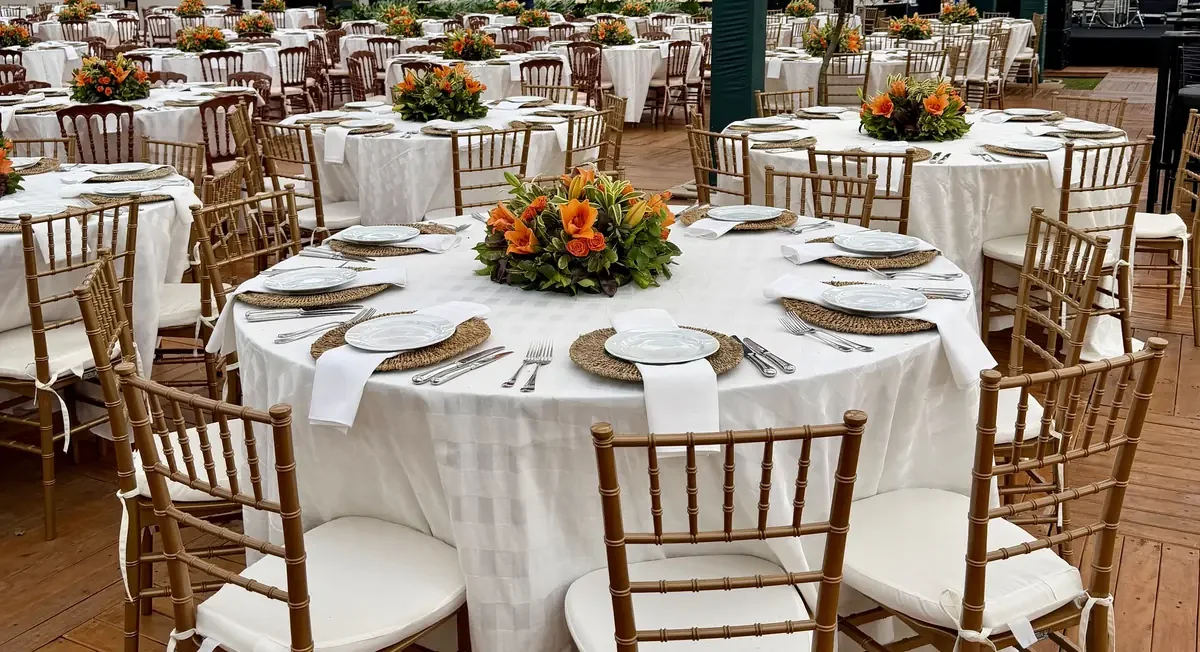 Mesas redondas montadas para jantar de empresa em pavilhão, com toalha branca, sousplats de fibra, guardanapos, arranjos centrais de flores laranja e cadeiras douradas de encosto ripado sobre piso de madeira