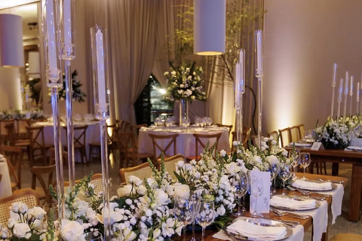Mesa longa de jantar posta em salão, com caminho de flores brancas e verdes no centro, castiçais altos de cristal, guardanapos com menu sobre os pratos e cadeiras de madeira ao fundo