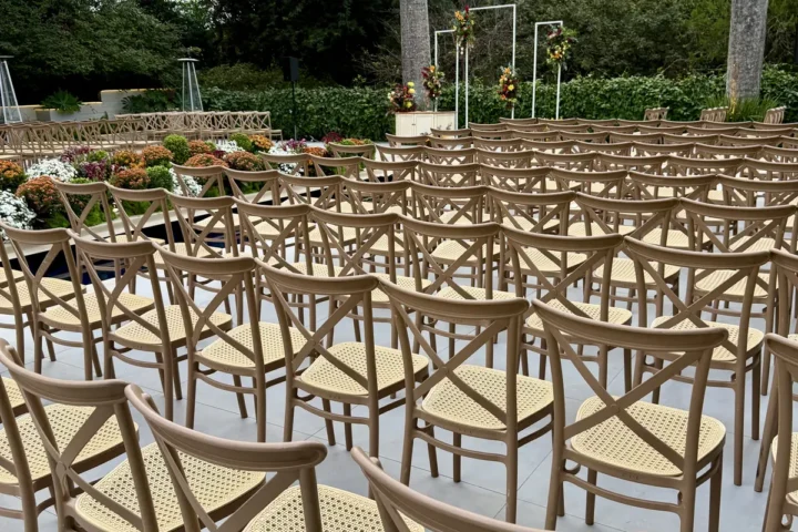 Fileiras de cadeiras de madeira com encosto cruzado e assento de palhinha, montadas em piso claro ao ar livre, diante de um arco de flores