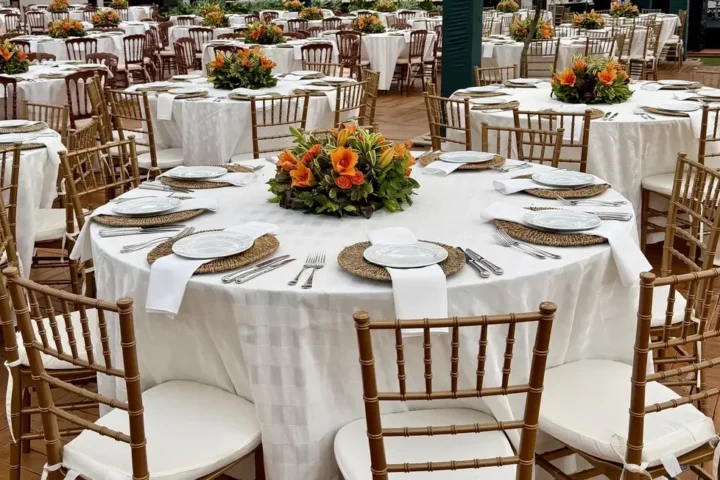 Mesas redondas montadas para jantar de empresa em pavilhão, com toalha branca, sousplats de fibra, arranjos de flores laranja e cadeiras douradas